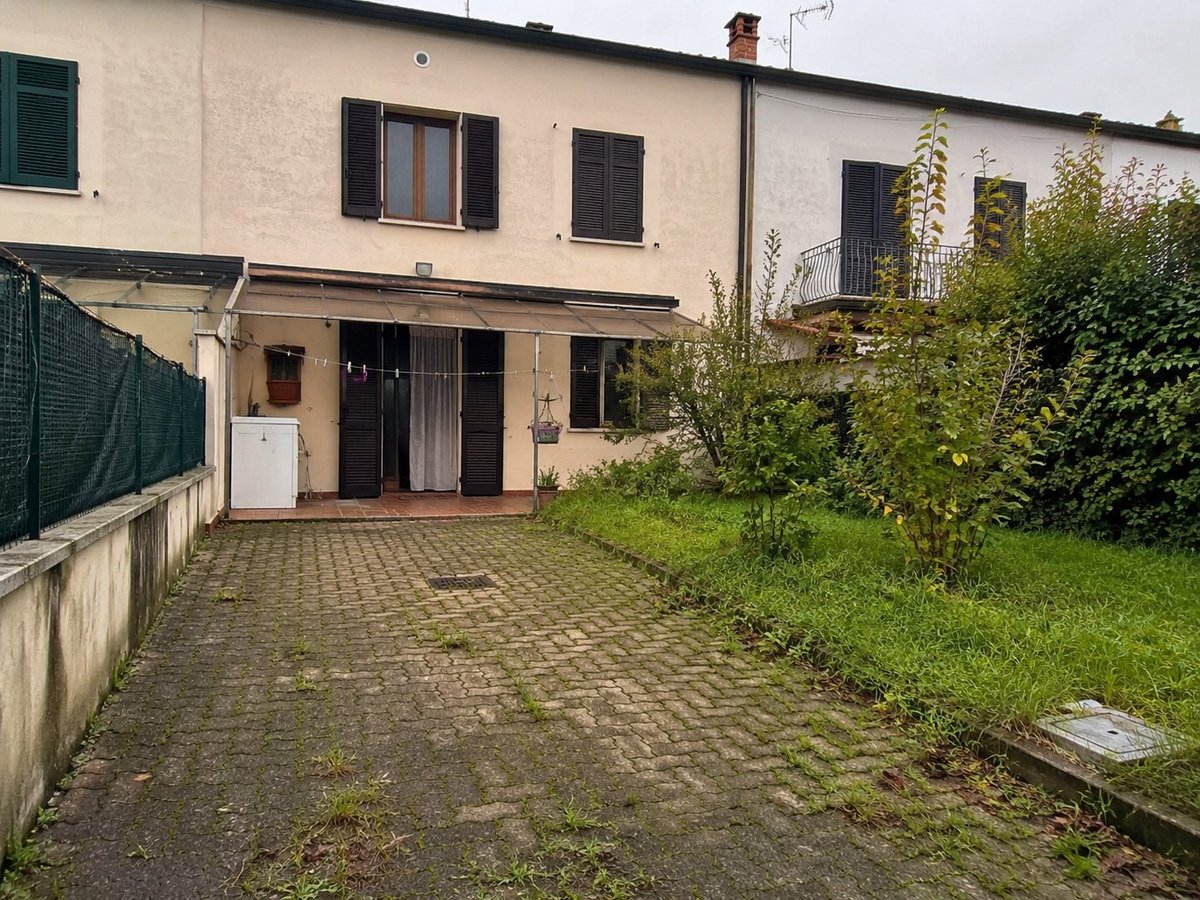 Single-story residential building with cream-colored facade, dark shutters, brick courtyard, and overgrown vegetation along the perimeter