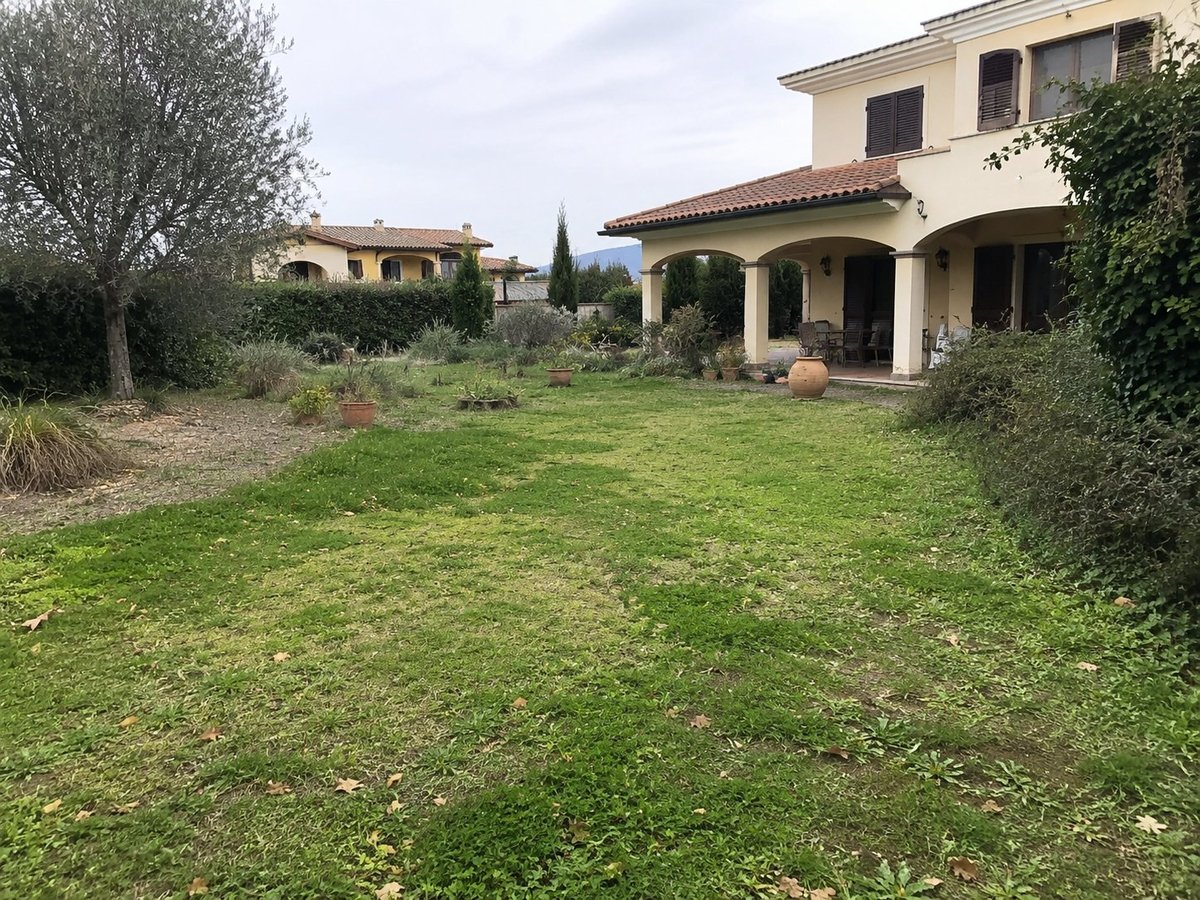 Mediterranean villa with cream-colored walls and arched portico, surrounded by green lawn and landscaping with olive trees and cypress.